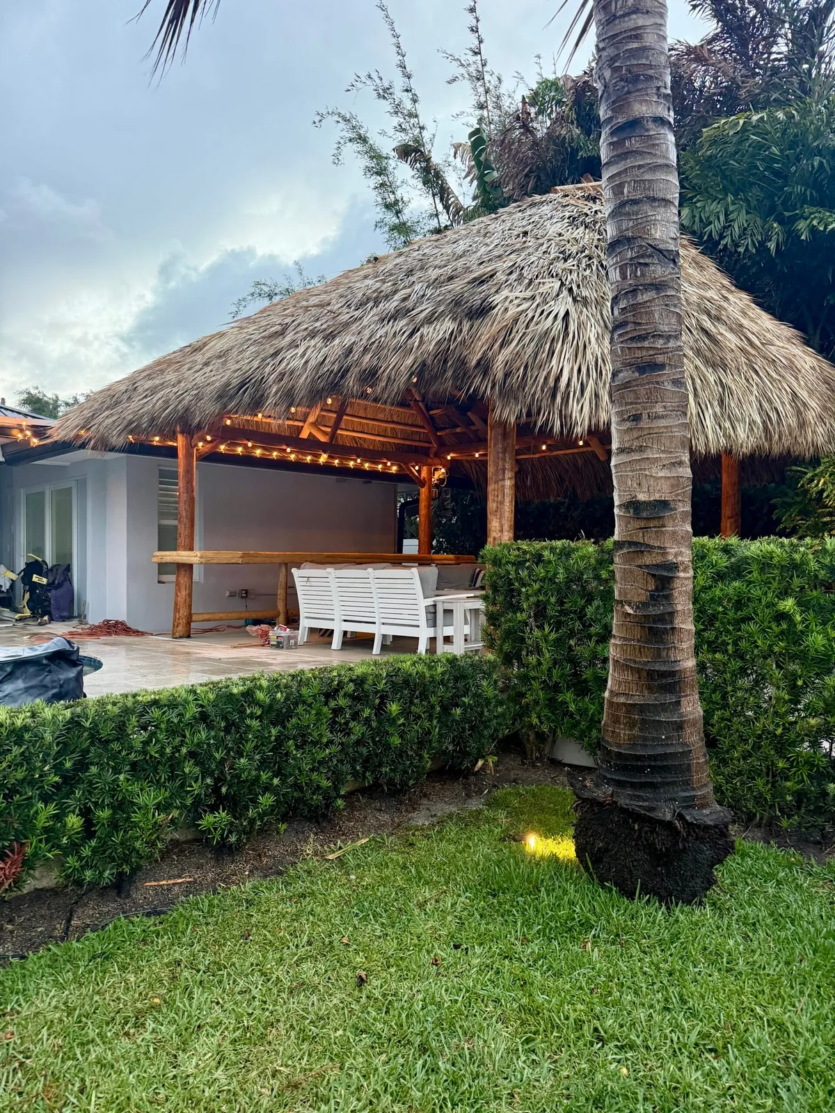 Natural palm tiki hut with warm string lights beside a backyard patio