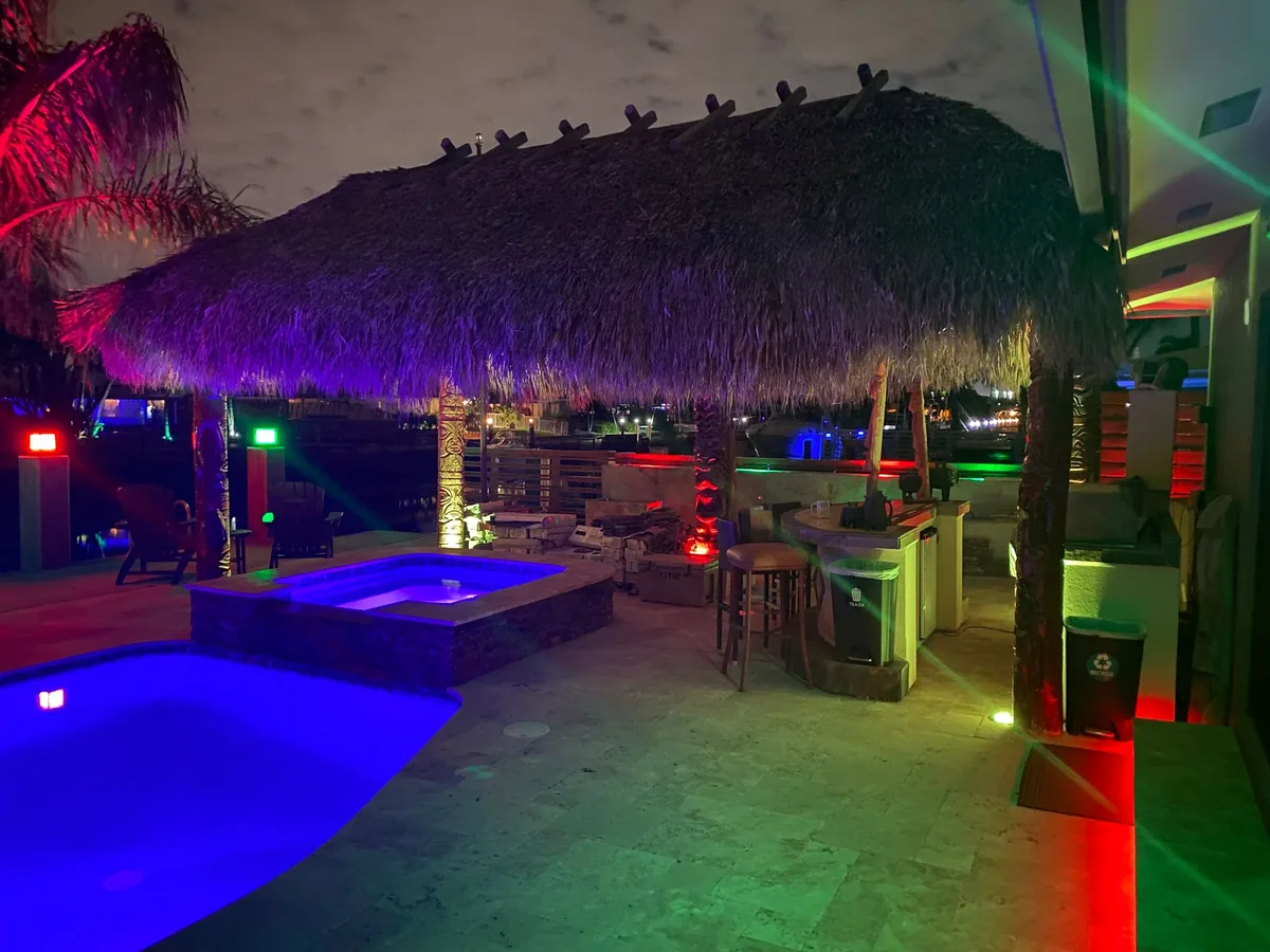 Tiki hut beside a swimming pool illuminated by colorful lights at night
